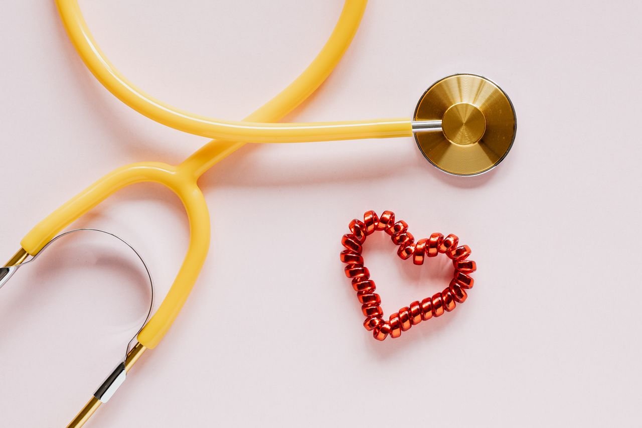 Top view of yellow stethoscope made of plastic and metal material near small handmade heart of spiral hair tie on pale pink background